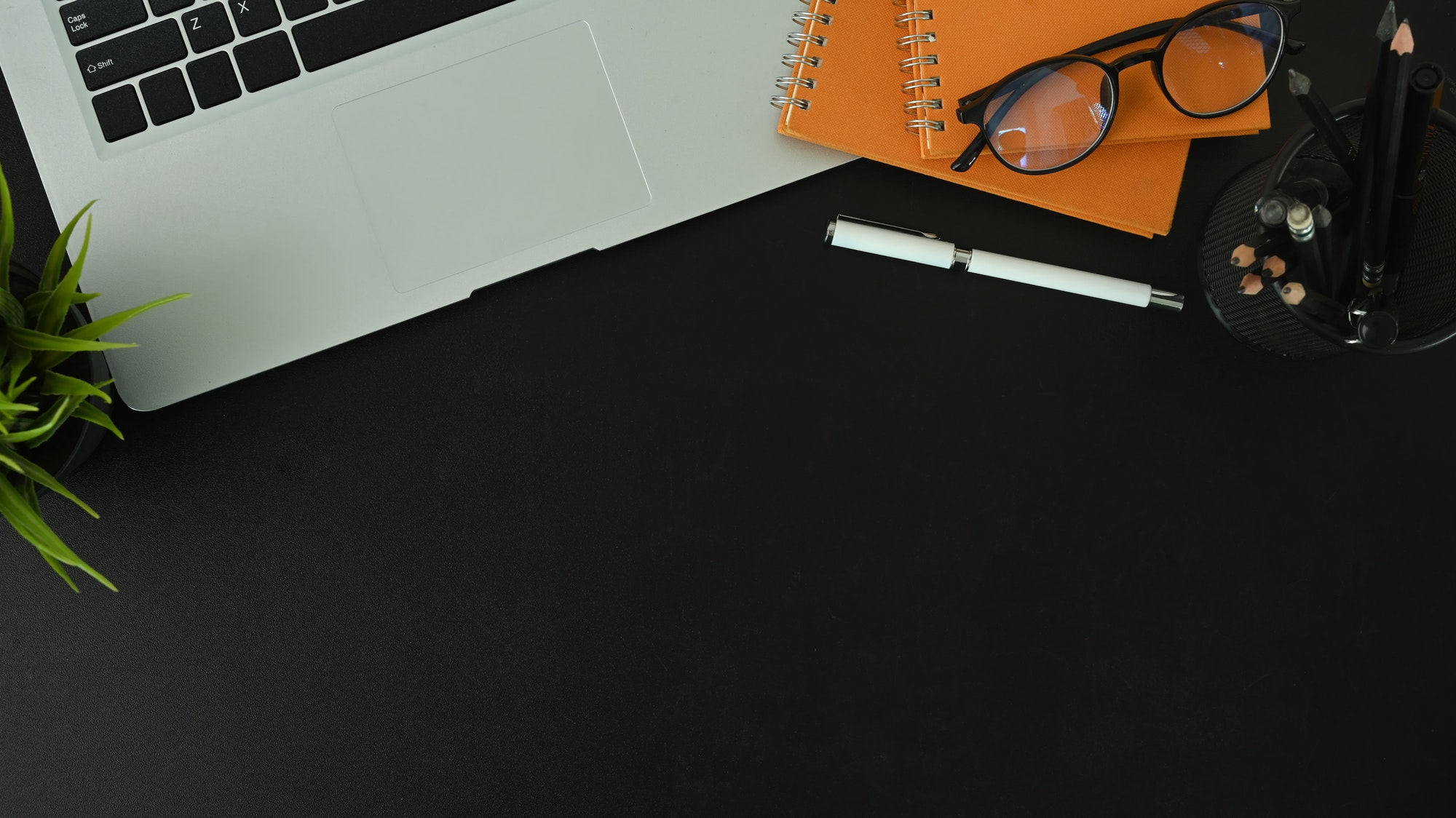 Dark office desk with laptop computer, pen, notebook glasses and plant on black table.